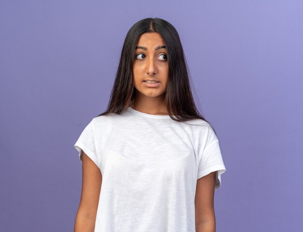 Chica joven en camiseta blanca mirando a un lado confundido y muy ansioso de pie sobre fondo azul.