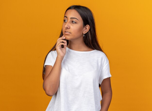 Chica joven en camiseta blanca mirando a un lado perplejo con expresión pensativa de pie sobre naranja