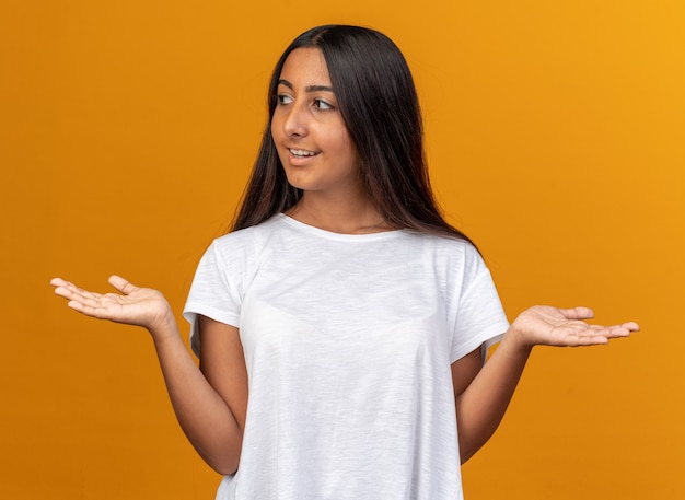 Chica joven en camiseta blanca mirando a un lado con una sonrisa en la cara extendiendo los brazos hacia los lados sobre naranja