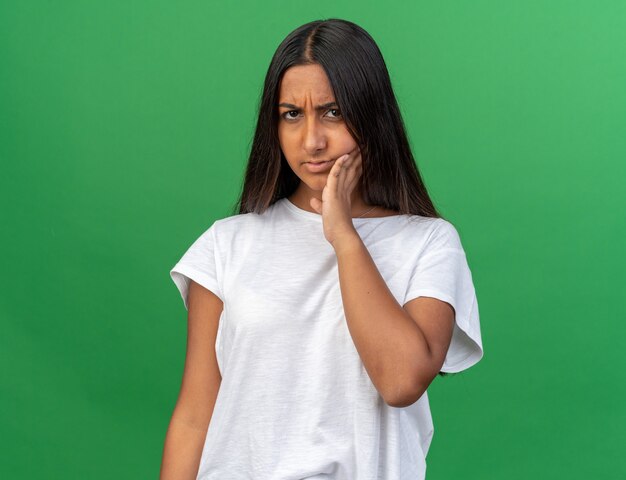 Chica joven en camiseta blanca mirando mal tocando su mejilla sintiendo dolor tener dolor de muelas de pie sobre fondo verde