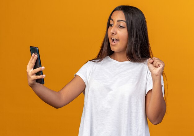 Chica joven en camiseta blanca mirando la pantalla de su smartphone apretando el puño feliz y emocionado de pie sobre fondo naranja