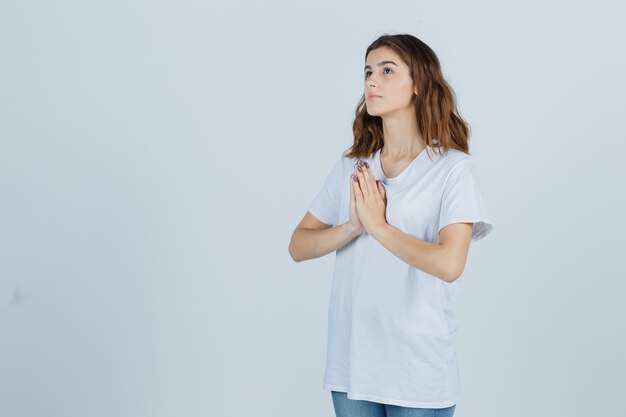 Chica joven en camiseta blanca mostrando gesto de namaste y mirando esperanzado, vista frontal.