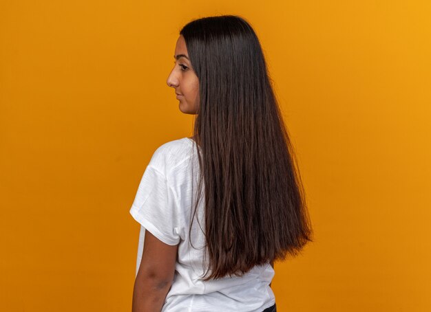 Chica joven en camiseta blanca de pie con la espalda sonriendo confiada sobre fondo naranja