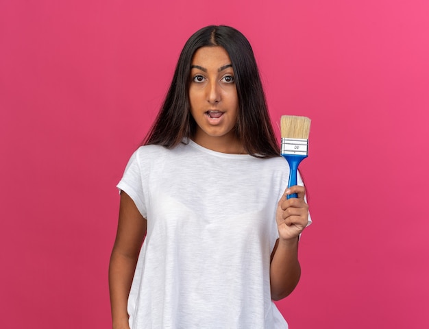 Chica joven en camiseta blanca con pincel mirando a cámara feliz y sorprendido de pie sobre rosa