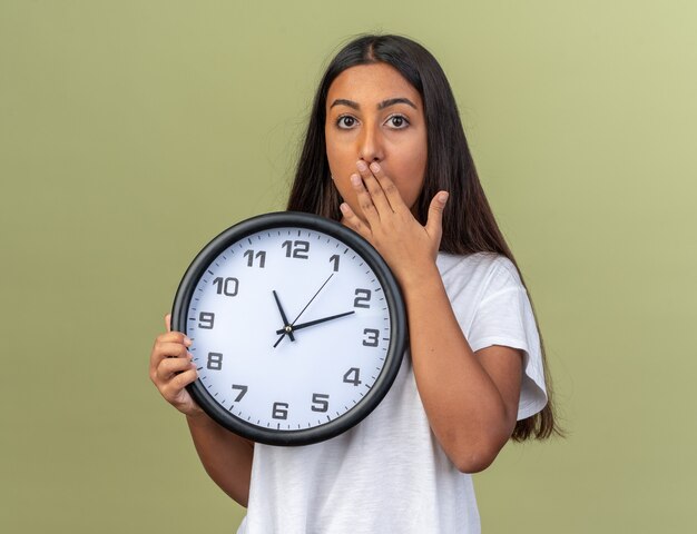 Chica joven en camiseta blanca con reloj de pared mirando a la cámara conmocionado cubriendo la boca con la mano