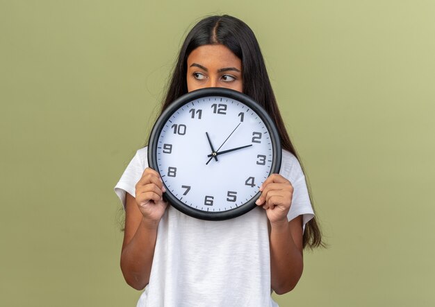 Chica joven en camiseta blanca con reloj de pared mirando a un lado con cara seria de pie sobre fondo verde