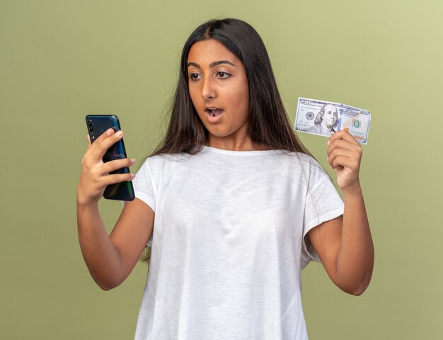 Chica joven en camiseta blanca con smartphone y dinero en efectivo mirando la pantalla de su móvil sorprendido y feliz
