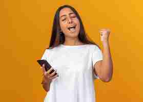 Foto gratuita chica joven en camiseta blanca con smartphone feliz y emocionado apretando el puño de pie sobre fondo naranja