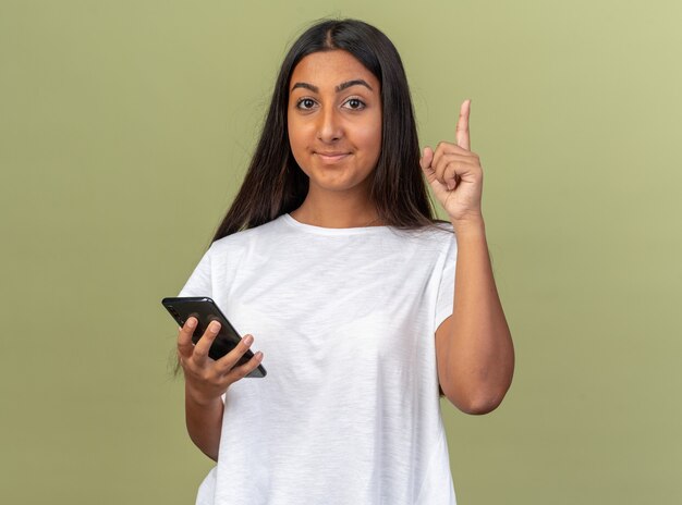 Chica joven en camiseta blanca con smartphone mirando a cámara con sonrisa en cara inteligente