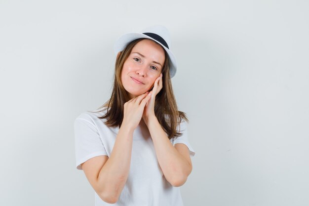 Chica joven en camiseta blanca, sombrero apoyado en la mejilla en la palma y mirando hermosa vista frontal.