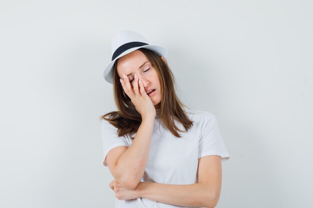 Chica joven con camiseta blanca, sombrero apoyando la mejilla en la palma levantada y mirando soñoliento, vista frontal.