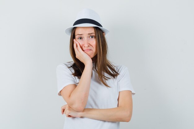 Chica joven en camiseta blanca, sombrero apoyando la mejilla en la palma y mirando triste, vista frontal.