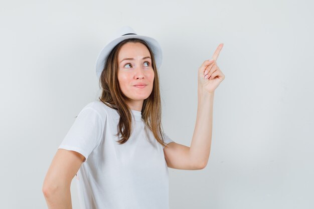 Chica joven con camiseta blanca, sombrero apuntando hacia arriba y mirando contento, vista frontal.