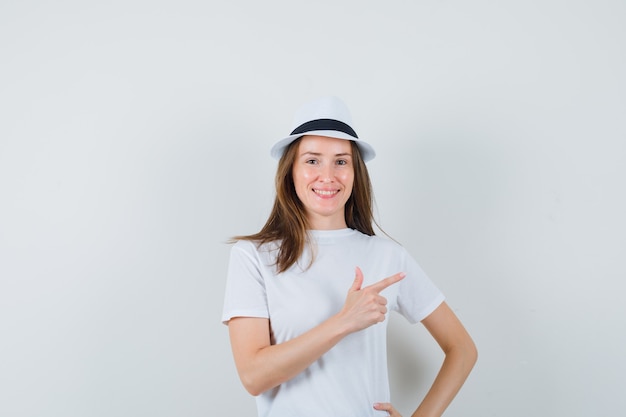 Chica joven con camiseta blanca, sombrero apuntando a la esquina superior derecha y mirando jovial, vista frontal.