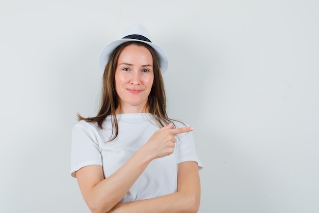 Chica joven en camiseta blanca, sombrero apuntando a un lado y mirando alegre, vista frontal.