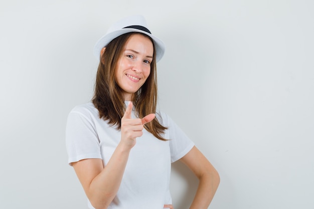 Chica joven en camiseta blanca, sombrero apuntando y mirando confiado, vista frontal.