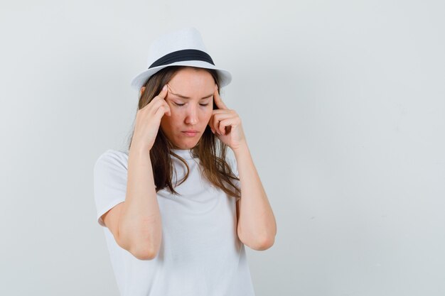 Chica joven en camiseta blanca, sombrero frotando sus sienes y mirando fatigado, vista frontal.