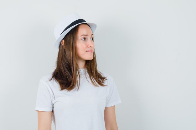 Chica joven con camiseta blanca, sombrero mirando a un lado y elegante vista frontal.