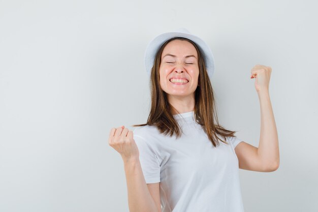 Chica joven en camiseta blanca, sombrero mostrando gesto de ganador y mirando feliz, vista frontal.