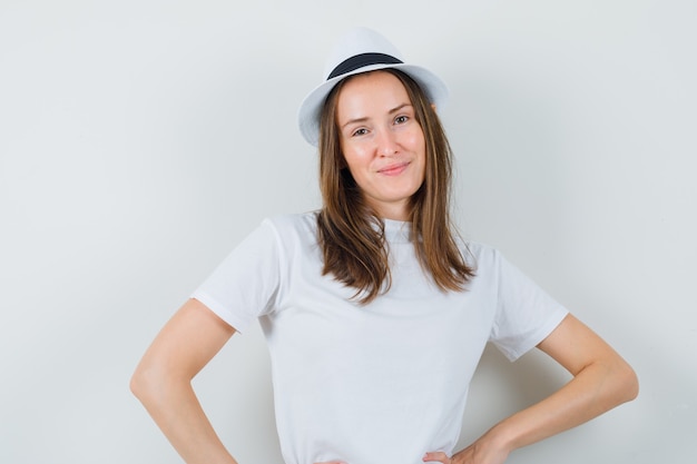 Chica joven en camiseta blanca, sombrero posando de pie y mirando glamorosa, vista frontal.