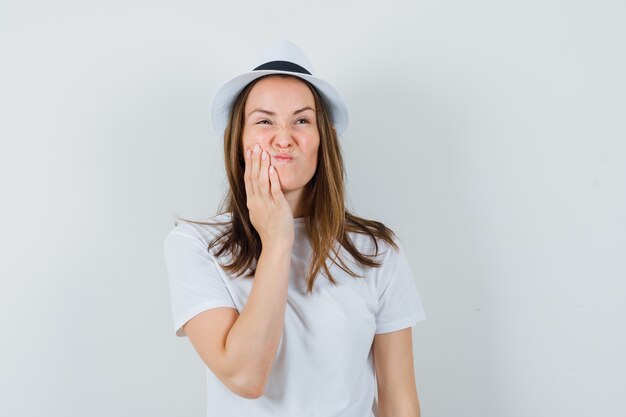 Chica joven con camiseta blanca, sombrero que sufre de dolor de muelas y se ve incómodo, vista frontal.