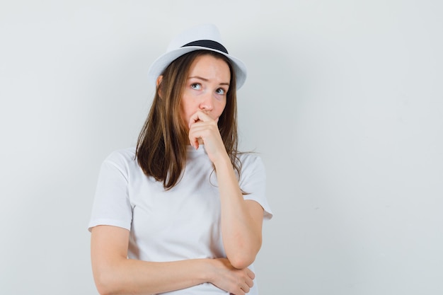 Chica joven con camiseta blanca, sombrero sosteniendo su barbilla y mirando pensativo, vista frontal.