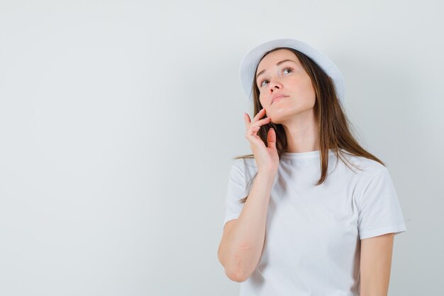 Chica joven con camiseta blanca, sombrero tocando la piel de la cara en la mejilla y mirando soñadora, vista frontal.