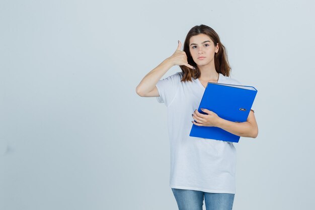 Chica joven en camiseta blanca sosteniendo una carpeta, mostrando el gesto del teléfono y luciendo útil, vista frontal.