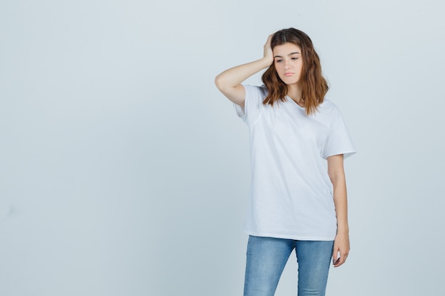 Chica joven en camiseta blanca sosteniendo la mano en la cabeza y mirando molesto, vista frontal.