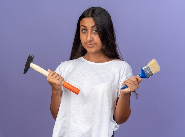 Foto gratuita chica joven en camiseta blanca sosteniendo un martillo y un pincel mirando a la cámara con una sonrisa de confianza en la cara de pie sobre azul