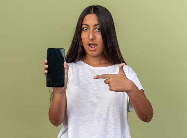 Chica joven en camiseta blanca sosteniendo smartphone apuntando con el dedo índice mirando confiado de pie sobre fondo verde