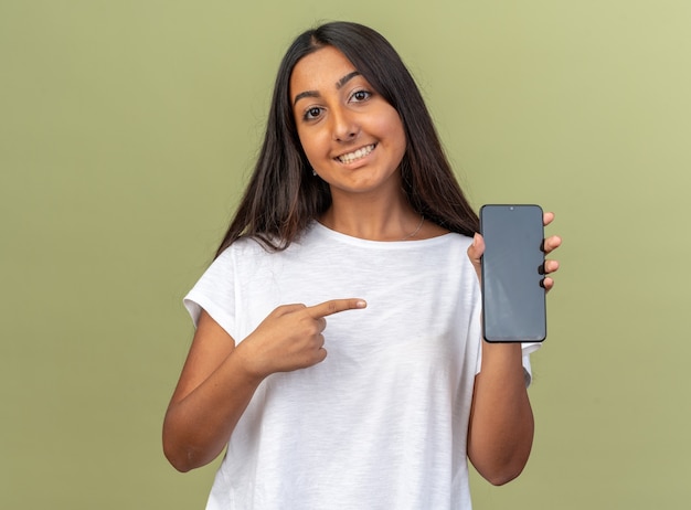 Chica joven en camiseta blanca sosteniendo smartphone apuntando con el dedo índice sonriendo alegremente