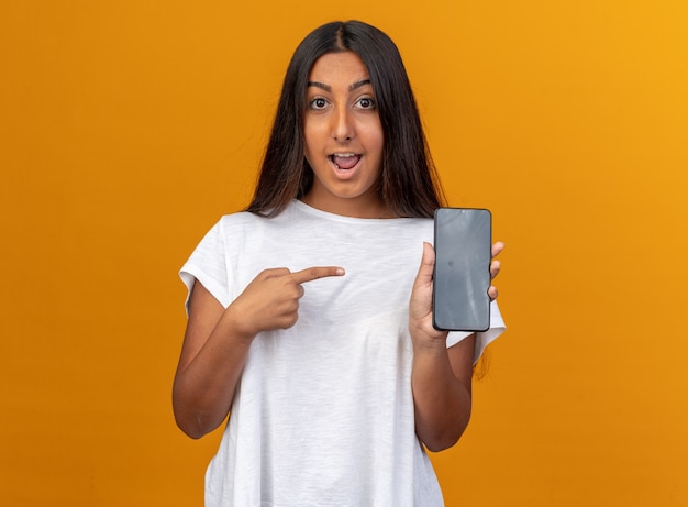 Chica joven en camiseta blanca sosteniendo smartphone apuntando con el dedo índice sonriendo