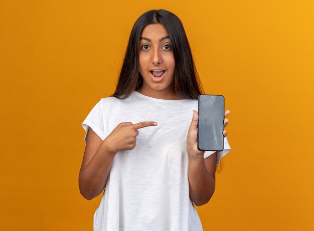 Chica joven en camiseta blanca sosteniendo smartphone apuntando con el dedo índice sonriendo