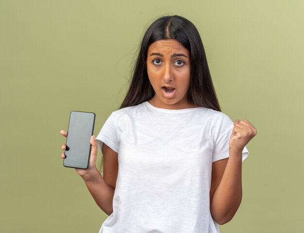 Chica joven en camiseta blanca sosteniendo smartphone mirando a cámara confundida y disgustada apretando el puño