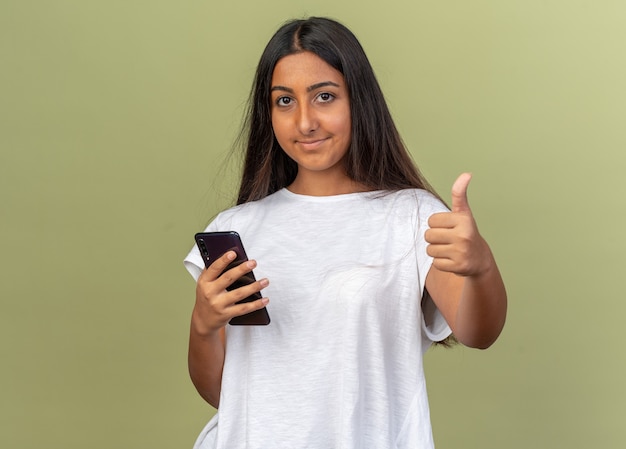 Chica joven en camiseta blanca sosteniendo smartphone mirando a la cámara sonriendo mostrando los pulgares para arriba sobre fondo verde