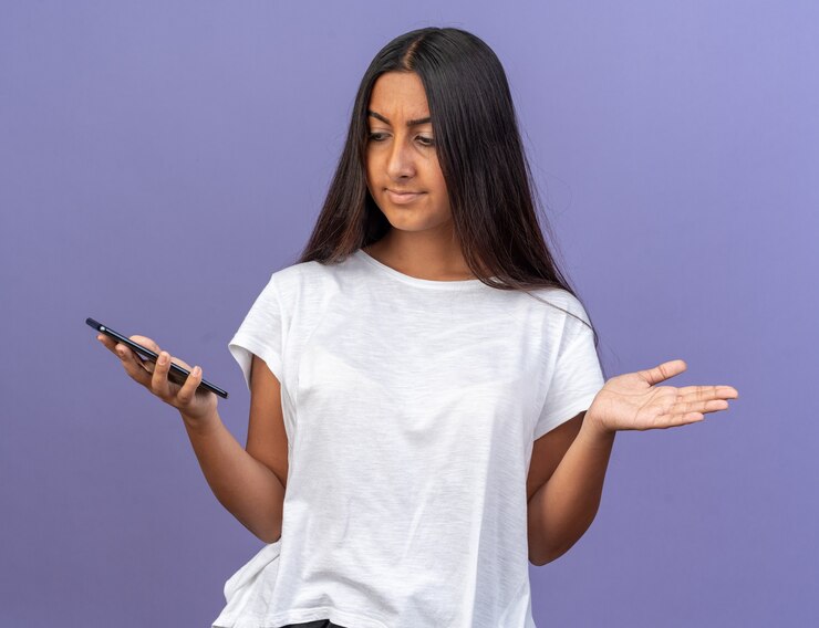 Foto gratuito chica joven en camiseta blanca sosteniendo smartphone mirándolo confundido extendiendo los brazos hacia los lados de pie sobre azul
