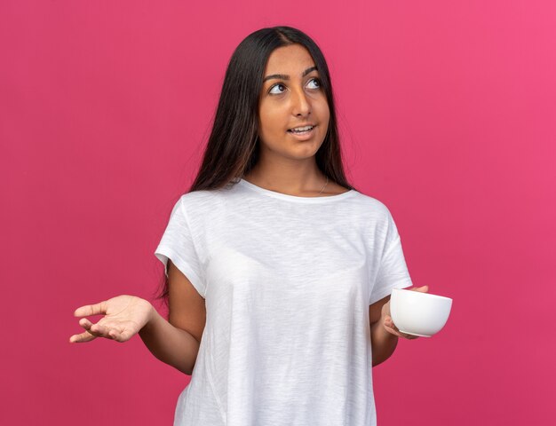 Chica joven en camiseta blanca sosteniendo una taza de café mirando hacia arriba con una sonrisa en la cara de pie sobre rosa