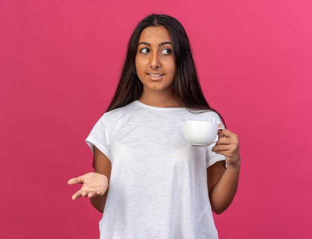 Chica joven en camiseta blanca sosteniendo una taza de té mirando a un lado con una sonrisa en la cara de pie sobre rosa