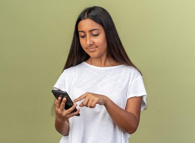 Chica joven en camiseta blanca sosteniendo un teléfono inteligente escribiendo un mensaje que parece seguro de pie sobre fondo verde