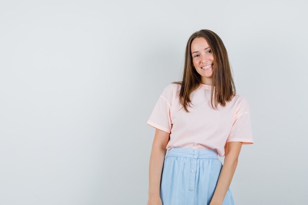 Chica joven en camiseta, falda mirando a cámara y mirando feliz, vista frontal.