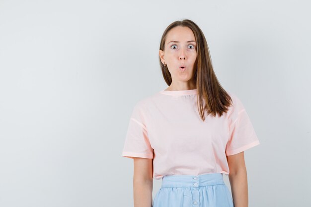 Chica joven en camiseta, falda mirando a cámara y mirando sorprendido, vista frontal.