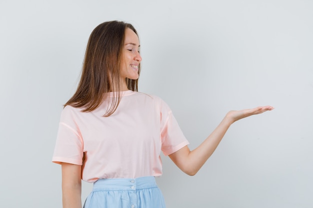 Chica joven en camiseta, falda mirando la palma extendida a un lado y mirando alegre, vista frontal.