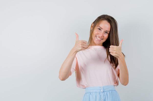 Chica joven en camiseta, falda mostrando los pulgares hacia arriba y mirando alegre, vista frontal.