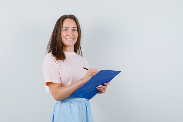 Chica joven en camiseta, falda tomando notas en el portapapeles y mirando alegre, vista frontal.