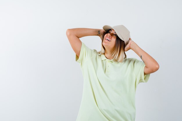 Chica joven en camiseta y gorra cogidos de la mano detrás de la cabeza, mirando a otro lado y mirando feliz, vista frontal.