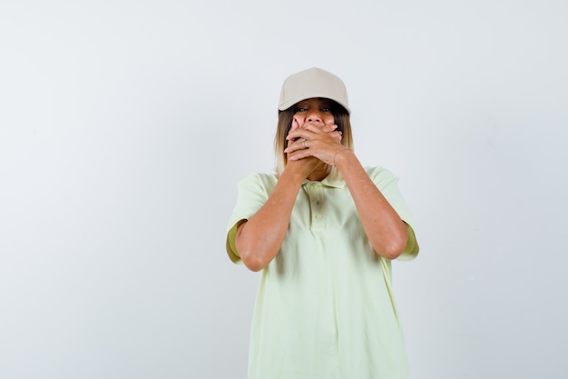 Chica joven en camiseta y gorra cubriendo la boca con las manos y mirando sorprendido, vista frontal.