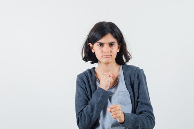 Chica joven en camiseta gris claro y sudadera con capucha frontal con cremallera gris oscuro apretando el puño mientras se señala a sí misma y se ve seria,