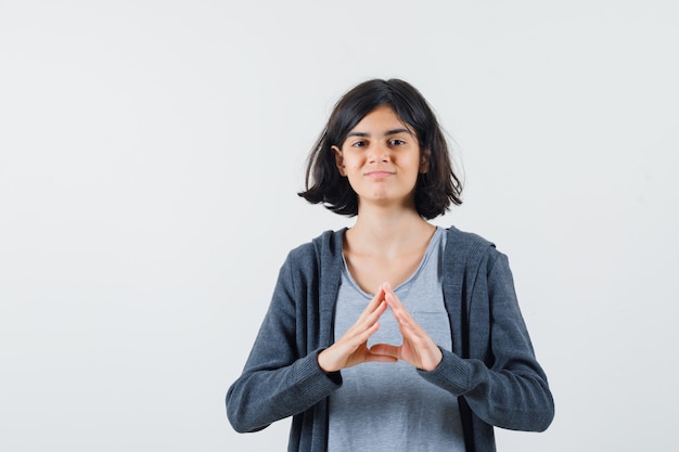 Chica joven en camiseta gris claro y sudadera con capucha frontal con cremallera gris oscuro que muestra forma de triángulo con las manos y se ve linda