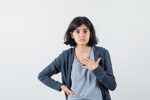 Chica joven en camiseta gris claro y sudadera con capucha frontal con cremallera gris oscuro sosteniendo la mano en la cintura, apuntando a sí misma y luciendo linda